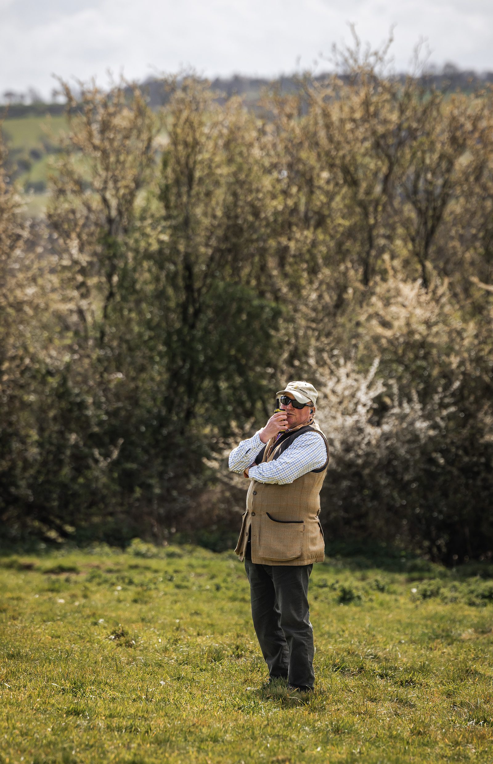A shoot host in tweed beside a blossoming spring hedgerow at Great Tew Estate in the Cotswolds