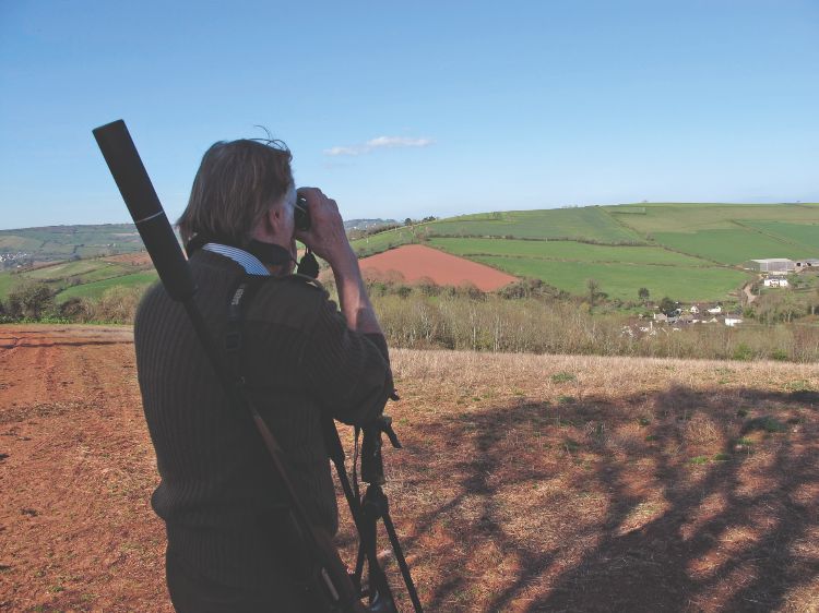 A man with a rifle on his back, scanning fields with binoculars